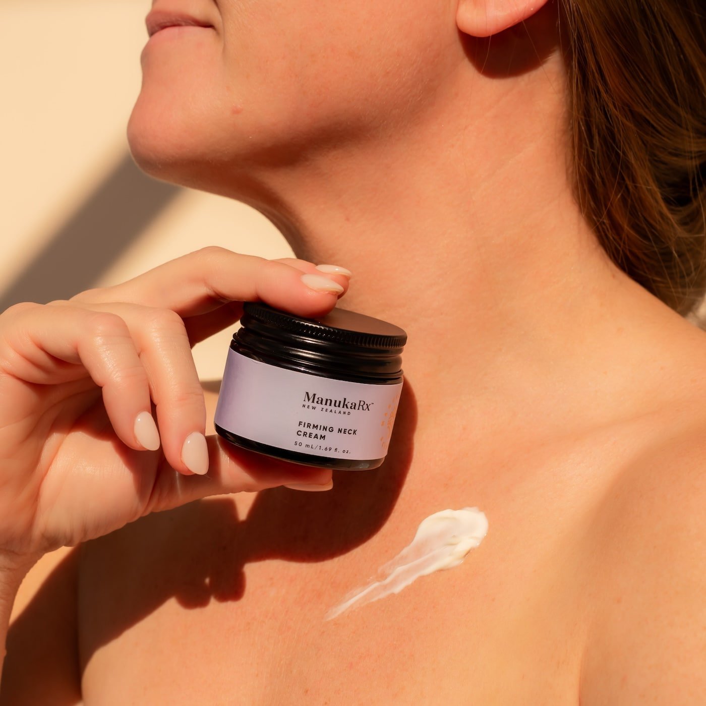 Woman holding jar of ManuakRx Firming Neck Cream with product swatch on her chest area